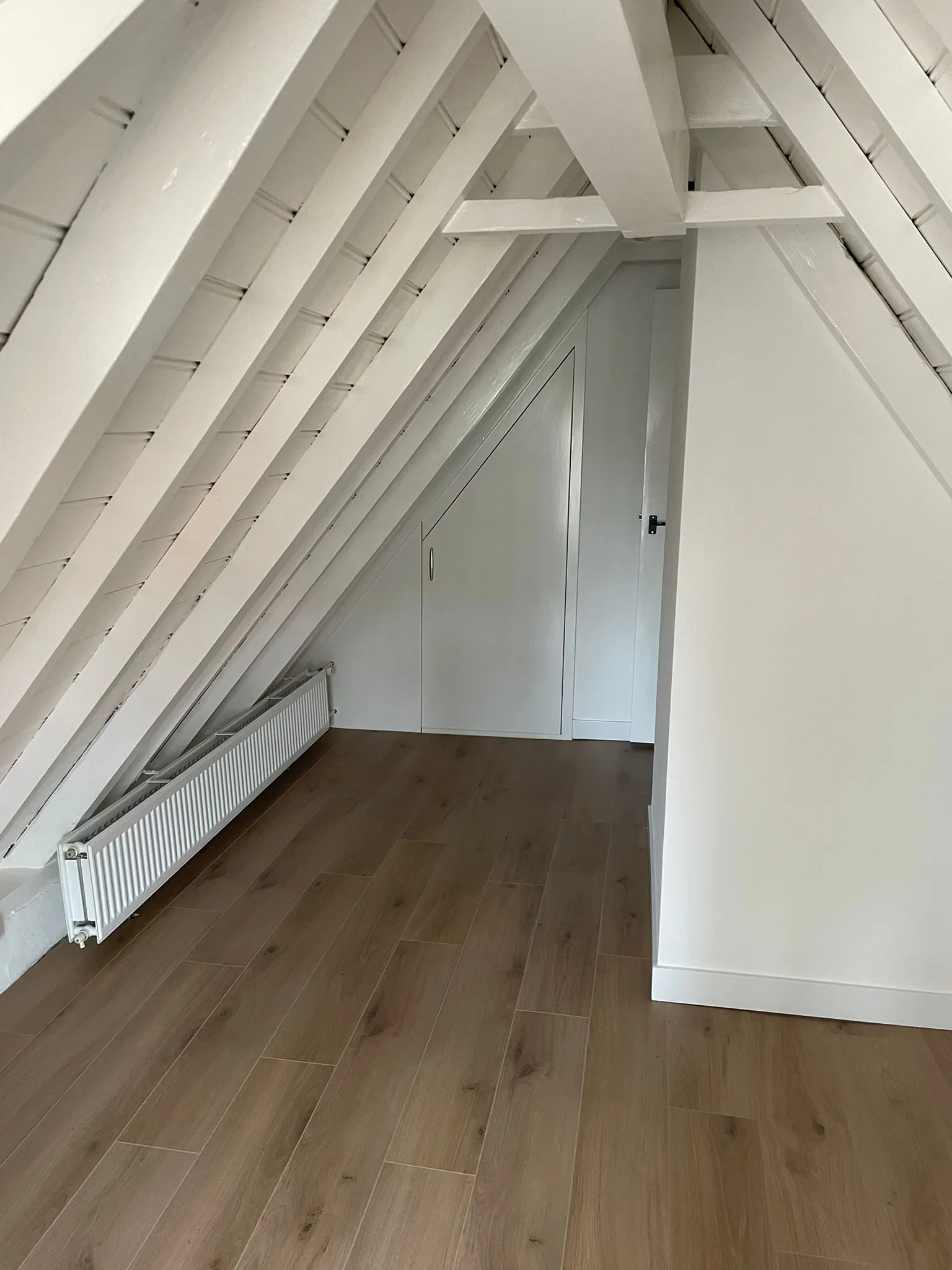 Zolderruimte met schuin dak, witte balken, houtlook vloer en een radiator langs de muur.